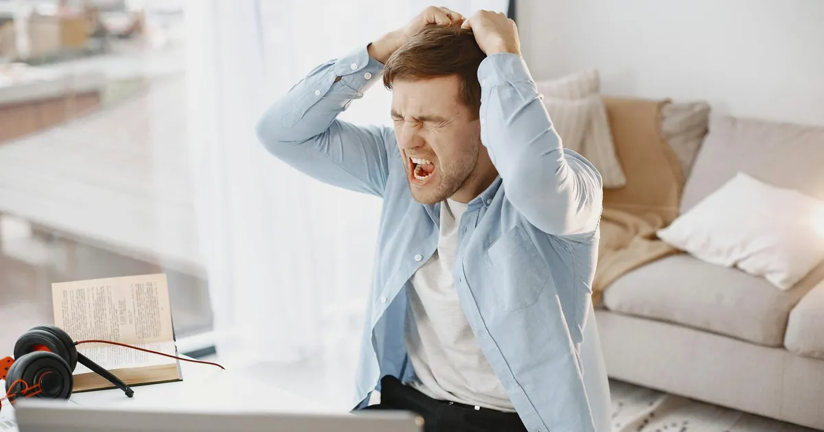 An upset man with mouth open, pulling hair at a desk with a laptop in a home setting.