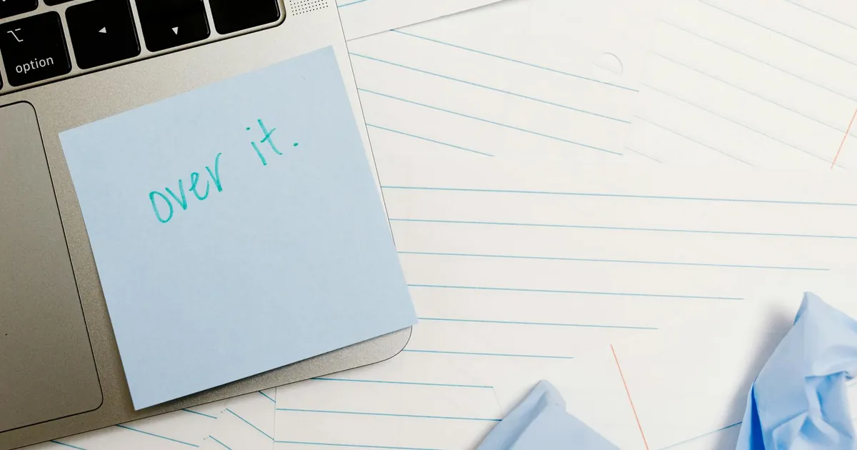 Close-up of laptop with sticky note saying 'over it' surrounded by lined papers and crumpled notes.