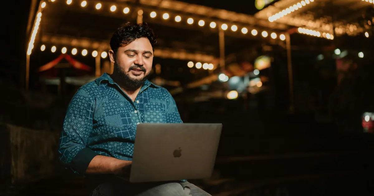 Adult man with a beard and mustache working on his laptop outdoors at night.