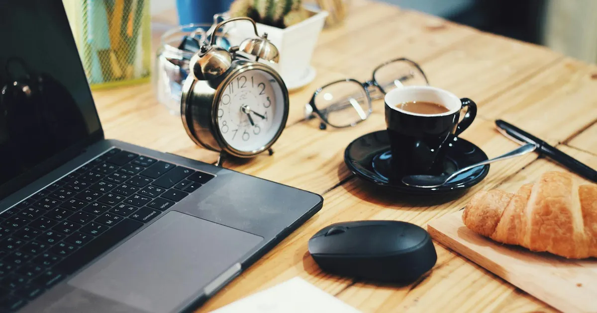 A warm workspace scene featuring a laptop, coffee, croissant, and clock on a wooden desk.