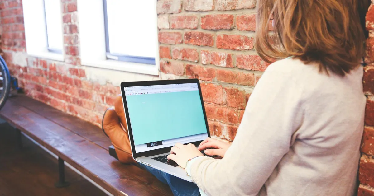 A woman is using a laptop for remote work while sitting indoors against a brick wall.