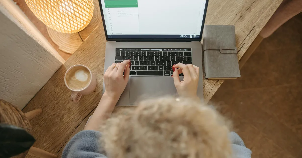 Overhead view of a person working on a laptop with coffee and journal on a wooden desk.