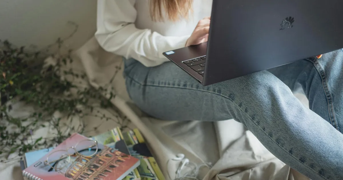 Crop anonymous woman in denim working on laptop sitting on bed with stack of books and notepads Crop anonymous woman in denim working on laptop sitting on bed with stack of books and notepads