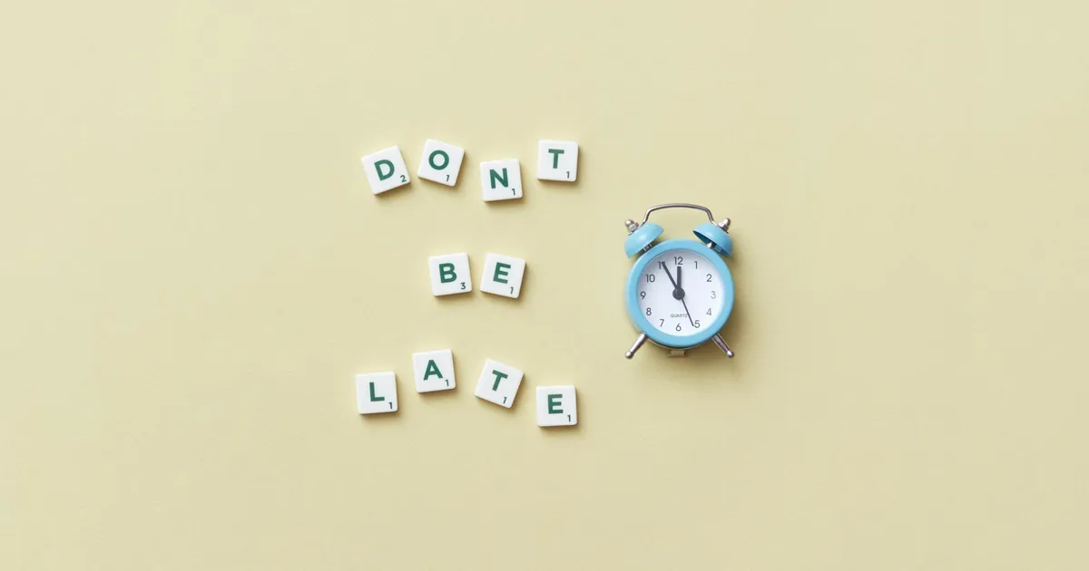 Flat lay of a blue alarm clock and letter tiles spelling 'Don't Be Late'.