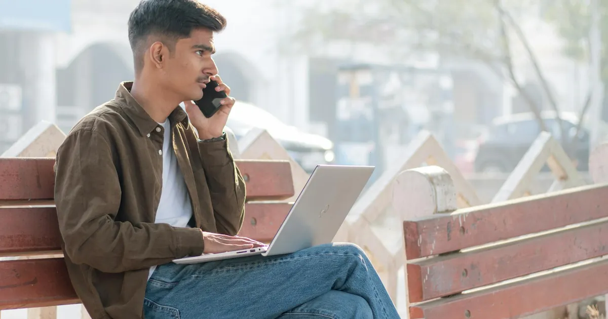 A young man multitasking with a laptop and smartphone on a park bench, embracing remote work outdoors.