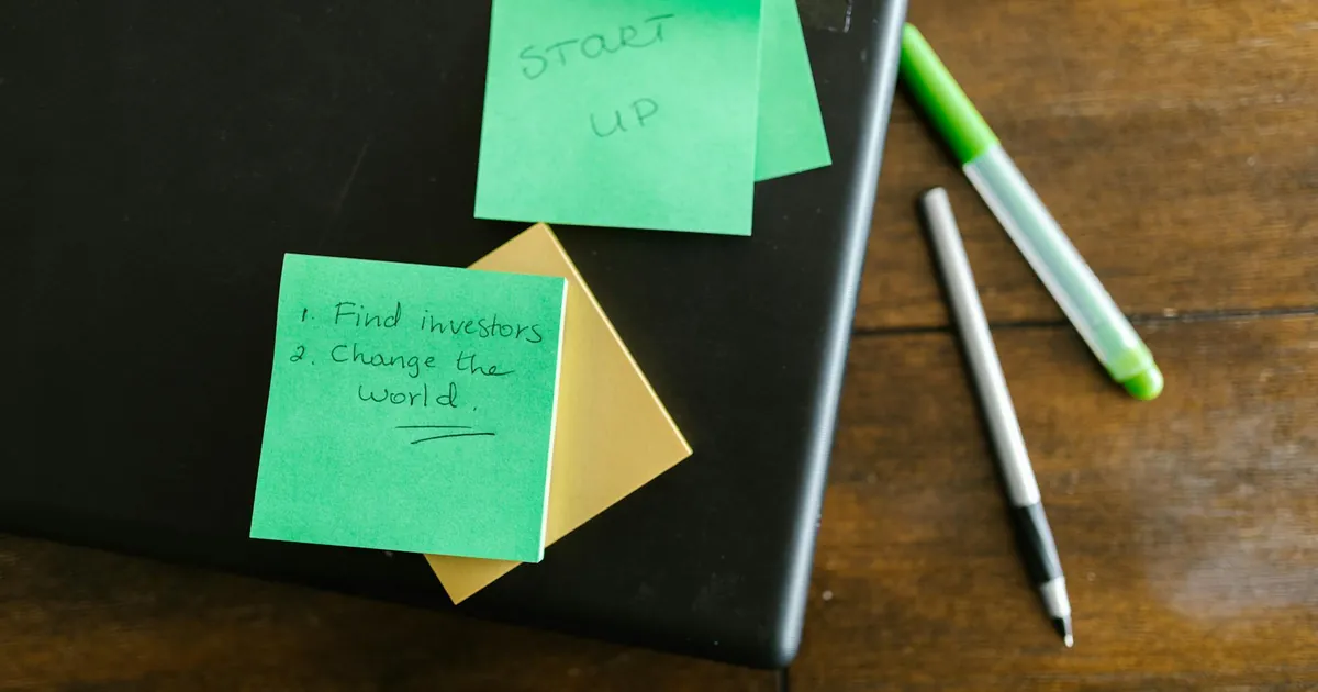 Green sticky notes with startup goals on a wooden desk with pens.