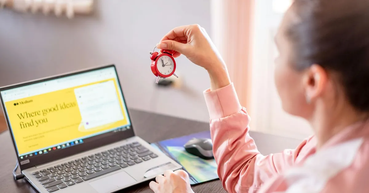 Woman holding a small clock at a desk with an open laptop screen displayed.