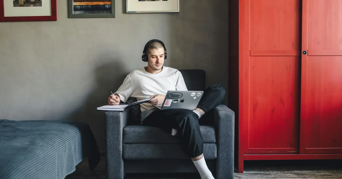 Man sitting in a cozy room working on a laptop with headphones, focusing on tasks.