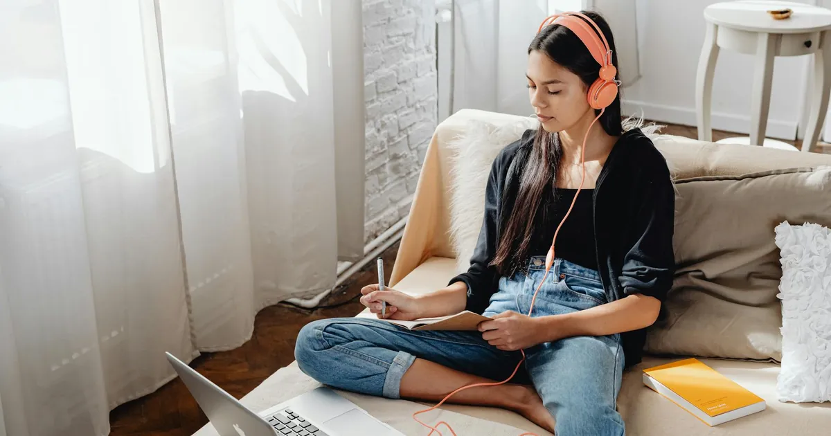 Young woman with headphones studying online from home on a sofa using a laptop.