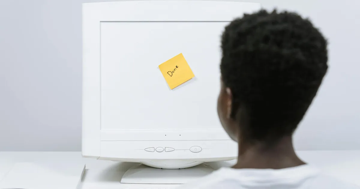 An unrecognizable person with afro hair looks at a monitor with a 'Done' sticky note, in a bright workspace. An unrecognizable person with afro hair looks at a monitor with a 'Done' sticky note, in a bright workspace.