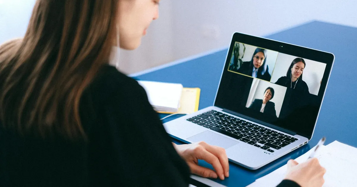 A woman engaging in a video conference using a laptop at home, taking notes.