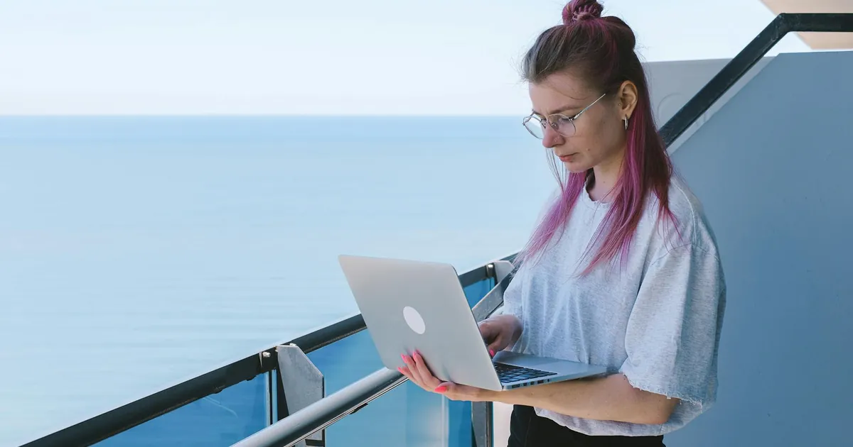 A young woman works remotely on her laptop by the sea on a sunny day, embodying the digital nomad lifestyle.