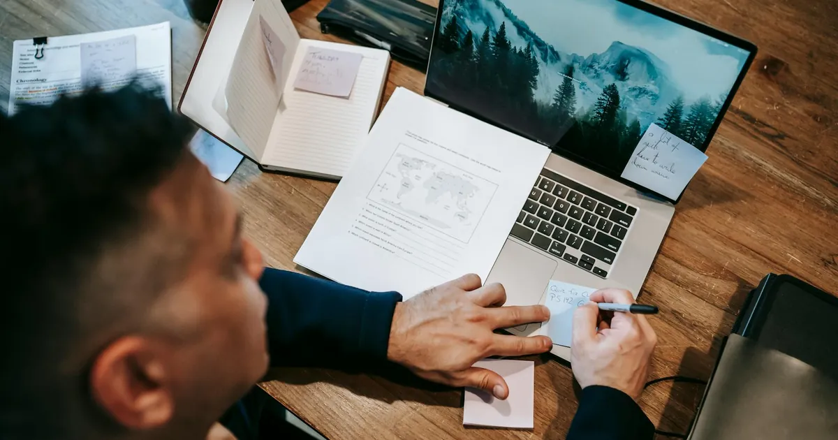 Man working remotely on laptop at a wooden desk with papers, notes, and a notebook.