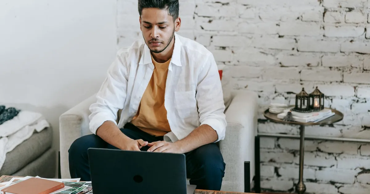 Creative young Hispanic male photographer sitting at table with photo camera and printed photos and working on laptop in modern loft style workplace at home