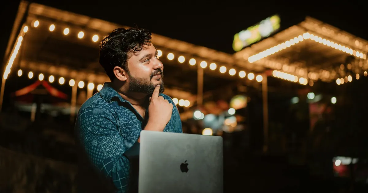 A thoughtful adult man with a beard using a laptop at night in a brightly lit outdoor setting.