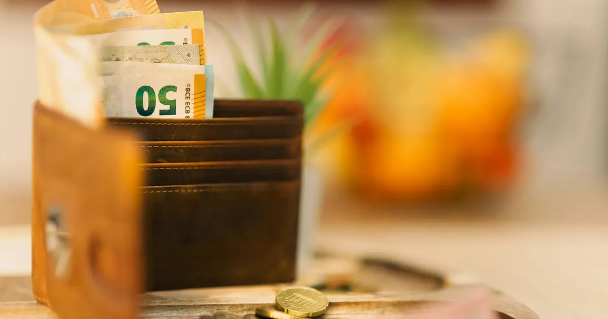 Brown wallet holding banknotes with scattered coins on a wooden table.