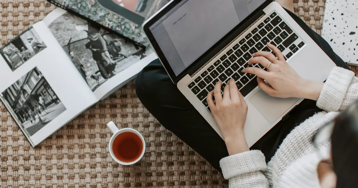 Overhead view of a woman typing on a laptop at home, surrounded by magazines and a cup of tea on the floor.