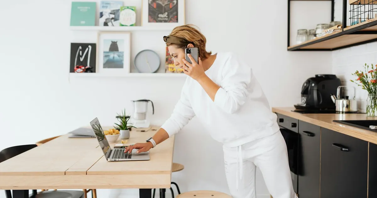 Woman using laptop and phone in stylish kitchen, blending work and home life.