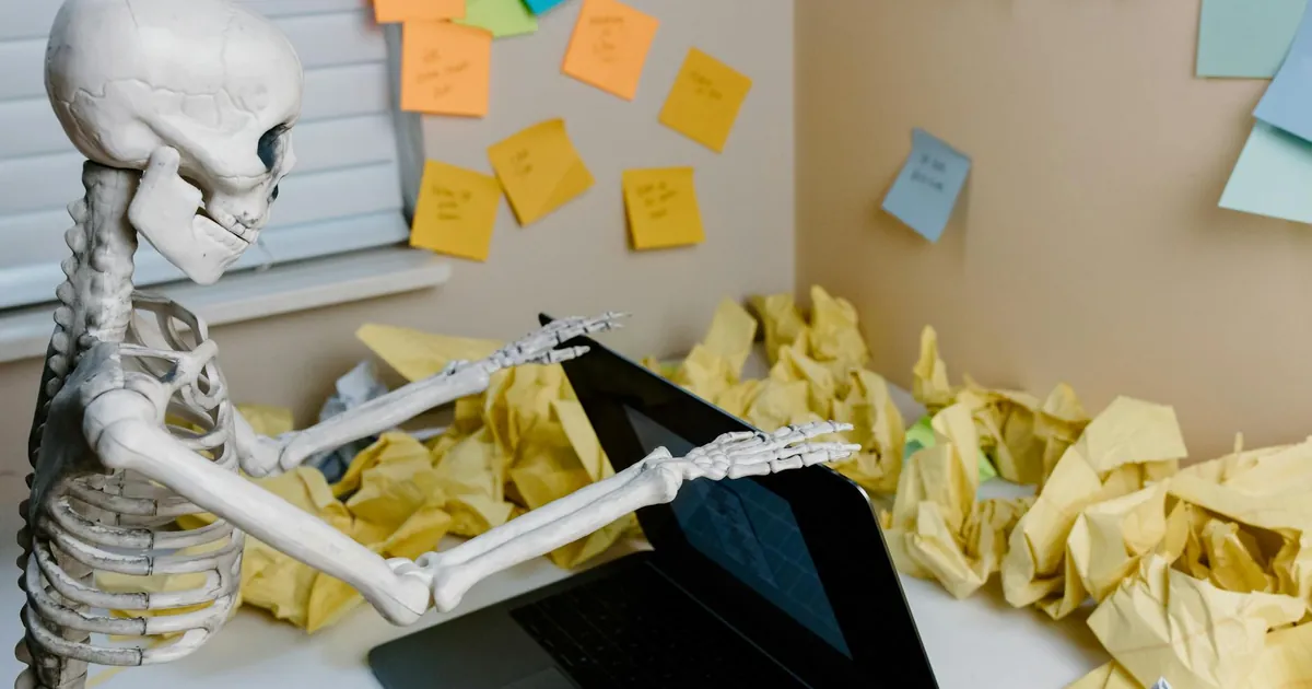 A skeletal figure works at a desk with a laptop and scattered papers, symbolizing burnout.
