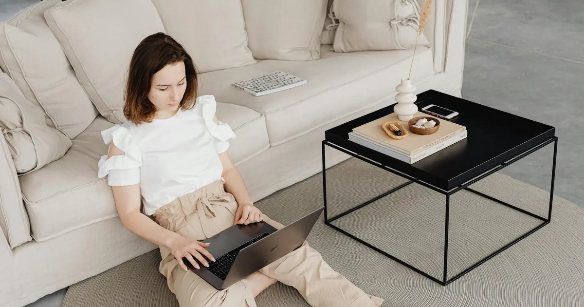 A woman sits comfortably on the floor working on her laptop in a modern living room.