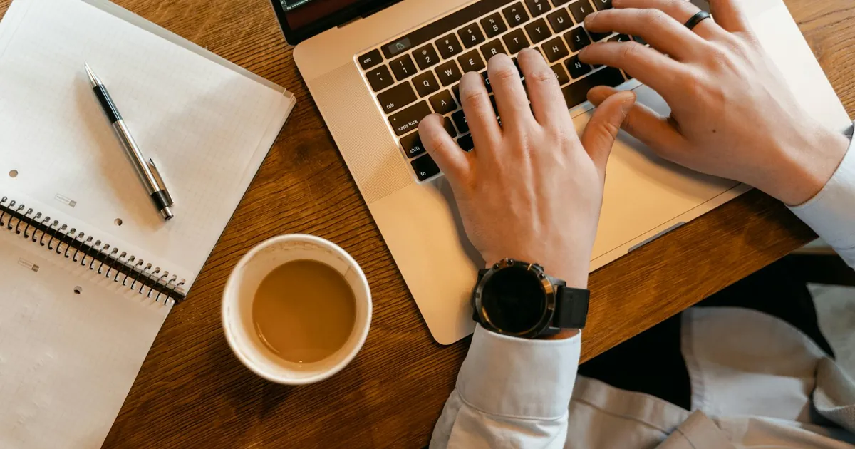 Hands typing on a laptop next to a cup of coffee and a notebook on a wooden table.