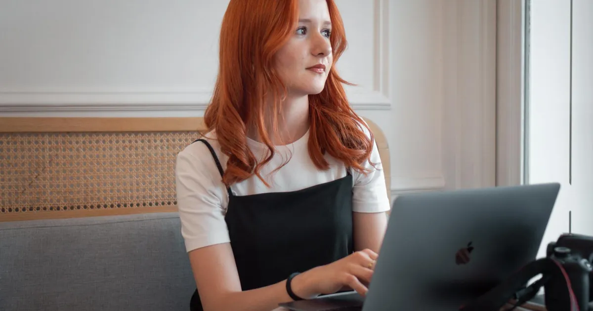 Young redhead woman working on laptop at a cafe, looking thoughtful.