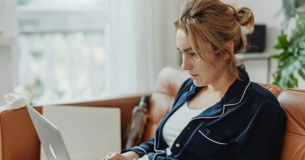 Woman in pajamas working on a laptop from a cozy home environment, focused on her task.