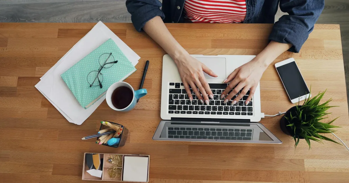 Top view of woman's hands typing on a laptop at a neatly organized wooden desk.